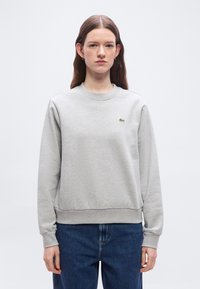 Jeune femme aux longs cheveux bruns portant un sweat-shirt gris clair à col rond avec un petit logo crocodile vert et un jean bleu, en position neutre.