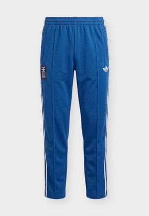 Siniset verkkarit, joissa valkoiset sivuraidat, joustava vyötärönauha, Adidas-logo oikeassa reidessä ja EMAX-tarra vasemmassa reidessä.