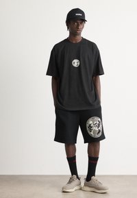 Mann iført svart Hugo-caps, oversized svart t-skjorte, svarte shorts med sirkulært logo, svarte sokker med rødt "Hugo" og beige sneakers som står mot en hvit vegg.