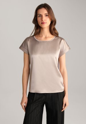 Silk-blend short-sleeve blouse in soft beige with a round neckline; smooth texture and relaxed fit. Paired with black pinstriped trousers.
