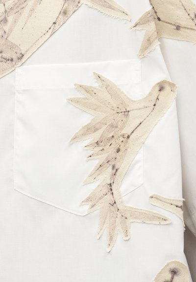 Λευκό πουκάμισο με μπροστινή τσέπη που παρουσιάζει μπεζ εφαρμογές πουλιών από ύφασμα με σκούρα κηλίδα στο φτερό και το σώμα.