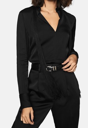 Femme portant un chemisier noir à manches longues et col en V, avec un pantalon noir assorti, une ceinture et un long accessoire à franges noires.