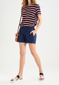 Žena v námořnických šortkách, námořnickém pruhovaném tričku s krátkým rukávem, stříbrných a černých sandálech a se hnědou taškou přes rameno.