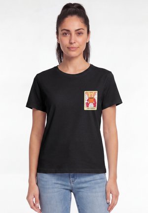 Femme aux cheveux foncés en queue de cheval portant un t-shirt noir avec un graphique coloré "The Coffee" sur la poitrine gauche et un jean bleu clair.