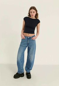 Svart croppet t-skjorte og lyseblå vide jeans med falmet finish, båret med kraftige svarte joggesko. Modellen står med hendene i lommene.