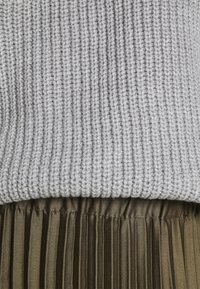Pull gris clair en maille superposé à une jupe plissée vert olive, montrant la texture et les détails du tissu.