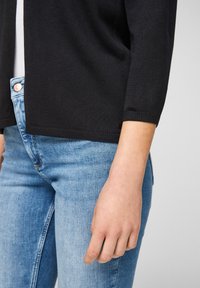 Black knitted cardigan with a round neckline, three-quarter sleeves, and a textured finish, paired with light blue denim jeans.