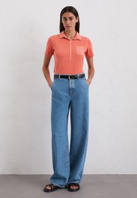 Koraalrood poloshirt met kraag en knoopsluiting, gecombineerd met hoge, wijde blauwe spijkerbroek en zwarte slide sandalen.