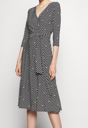 Frau trägt ein schwarz-weißes Wickelkleid mit geometrischem Muster, dreiviertel Ärmeln und einem Bindegürtel an der Taille.