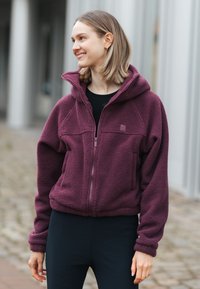 Nainen hymyilee ja katsoo sivulle, päällään lämmin violetti vetoketjullinen fleece-takki ja mustat housut ulkona asfaltoidulla polulla.