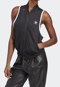 Černá bezrukávová bunda na zip s vysokým límcem, bílými akcenty a bočními kapsami. Na hrudi je logo Adidas a bunda má hladkou texturu.