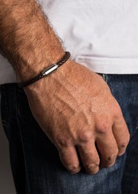 Bracciale in pelle intrecciata marrone con chiusura cilindrica metallica, indossato su un polso, che mette in mostra fili testurizzati e un accento metallico liscio.