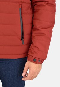 La chaqueta acolchada roja presenta un bolsillo con cremallera en el pecho y un puño con botón a presión. La tela es suave con secciones acolchadas.