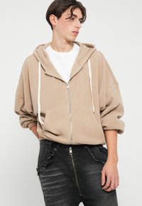 Sweatshirt bege com fecho éclair, de textura suave, capuz grande e cordões brancos. Combinado com jeans escuros desgastados com fecho na parte da frente.