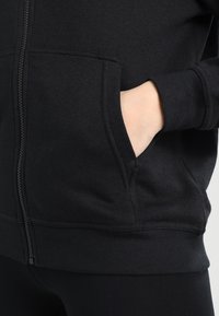 Hand inside the front pocket of a black zip-up hoodie worn with black pants, showing fabric texture and stitching details.