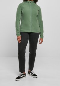 Giacca in pile verde, con zip frontale, design con cappuccio, maniche raglan, tasche laterali, abbinata a jeans scuri e sneakers nere.