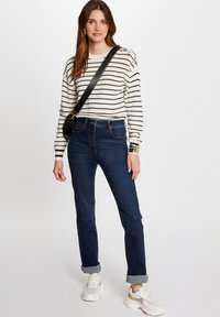 Suéter de rayas en crema y negro, jeans azul oscuro de corte ajustado y zapatillas blancas. Presenta botones dorados en el hombro del suéter.