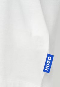 Λευκό βαμβακερό μπλουζάκι με μπλε υφασμάτινη ετικέτα που αναγράφει "HUGO" με λευκά γράμματα. Απλός σχεδιασμός, απαλή υφή και κλασικό σχήμα.