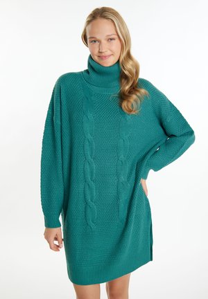 Jeune femme aux longs cheveux blonds portant une robe-pull en maille torsadée à col roulé bleu sarcelle, debout, une main sur la hanche.