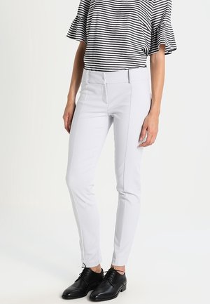 Kobieta ubrana w białe, dopasowane spodnie, czarne skórzane oksfordy oraz czarno-białą, paskowaną bluzkę z falbankami na półrękawkach.