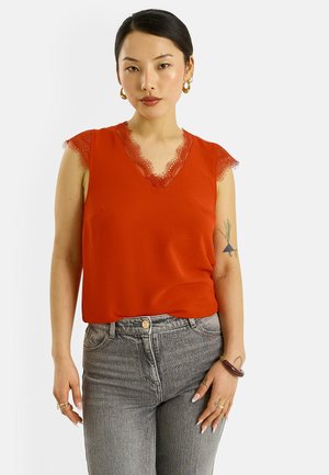 Femme aux cheveux noirs portant un haut rouge sans manches avec bordure en dentelle et un jean gris, debout les mains détendues le long du corps.