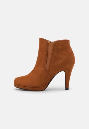 Brun ruskinds ankelstøvle med elastisk panel i siden og høj stiletthæl på hvid baggrund.