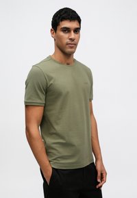 Mladý muž s krátkymi tmavými vlasmi, oblečený v olivovo zelenom tričku s krátkym rukávom a čiernych nohaviciach, stojaci s jednou rukou v vrecku.