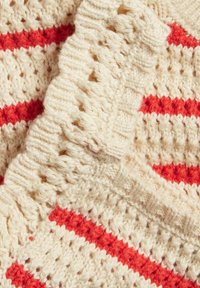 Häggt textil med en krämfärgad bas och röda ränder, med ett texturerat mönster och scallopade kanter för extra detalj.