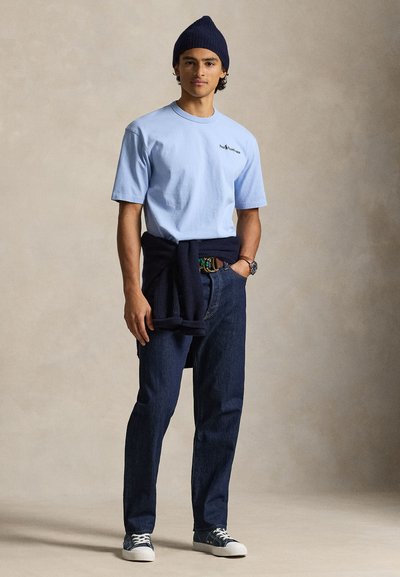 Ανοιχτό μπλε βαμβακερό t-shirt, σκούφος πλεκτός σε ναυτικό μπλε, σκούρα μπλε τζιν παντελόνια και αθλητικά παπούτσια. Πουλόβερ δεμένο στη μέση, με λεπτομέρεια στη ζώνη ορατή.