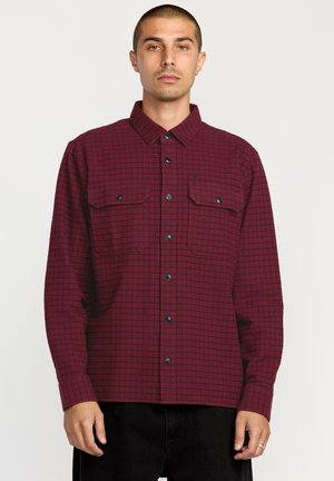 Red and black checkered long-sleeve shirt with two chest pockets and button closure, made of textured fabric, displayed on a model.