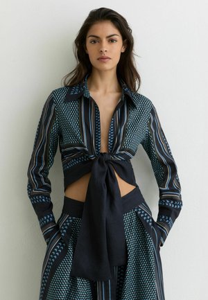 Femme aux longs cheveux foncés portant un crop top noué à motifs noirs et bleus et un pantalon taille haute assorti, debout contre un mur blanc.