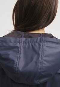 Långt mörkt hår som faller över en marinblå vattentät jacka med huva, bärs av en person som står vänd bort från kameran.