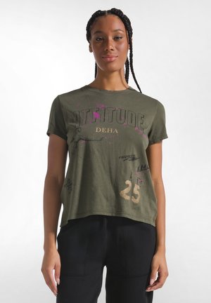 Donna con i capelli intrecciati che indossa una maglietta verde oliva con le parole attitude, DEHA e il numero 25, abbinata a pantaloni neri, in piedi su sfondo bianco.