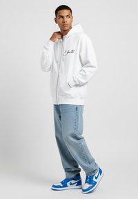 Felpa bianca con zip e testo stampato, con tasche frontali, abbinata a jeans azzurri e sneakers bianche/blu. Tessuto morbido, vestibilità comoda.
