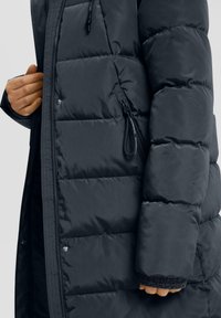 Tamsus paminkštintas žieminis paltas su užtrauktuku ir spaudėmis, viena ranka reguliuojanti paltą, matomas pūkuotas rankovės ir užtrauktuko kišenių detalės.