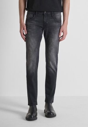 Pantalones vaqueros de mezclilla negra con corte ajustado, lavado deslavado, cinco bolsillos y detalles de botón de metal, combinados con botas negras.