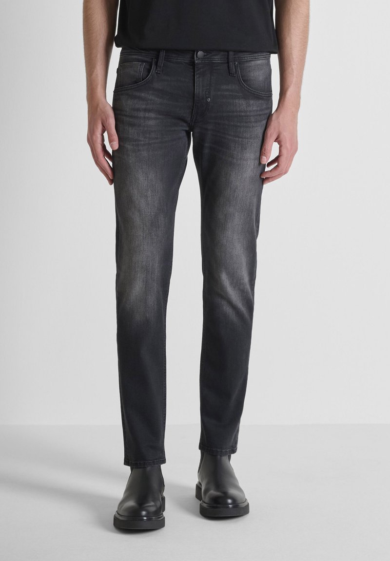 Pantalones vaqueros de mezclilla negra con corte ajustado, lavado deslavado, cinco bolsillos y detalles de botón de metal, combinados con botas negras.