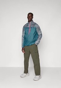 Άνδρας φοράει τζάκετ σε τιρκουάζ και γκρίζο χρώμα με μισό φερμουάρ, ελαιόχρωμα παντελόνια cargo και λευκά αθλητικά παπούτσια, μπροστά σε απλό αχνό φόντο.