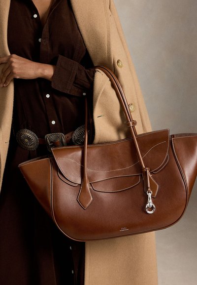 Person trägt ein braunes Hemd und einen beigen Mantel und hält eine große braune Ledertasche mit doppelten Griffen und silberner Schnalle.