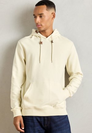Sudadera con capucha de color crema, de textura acanalada, con cordones ajustables y bolsillo tipo canguro. Puños y cinturilla ajustados.