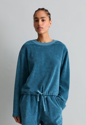 Kobieta z warkoczami, ubrana w turkusowo-niebieską bluzę z troczkami i dopasowane spodnie, stojąca na jednolitym szarym tle.