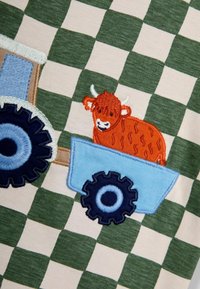 Vaatteeseen broderad bomull med ett grönt och beiget rutmönster, en blå traktor med mörka hjul och en orange ko i en släpvagn.