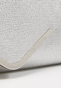 Zilveren clutch met textuur, voorzien van een metalen accent en logo-detail. Heeft een slank ontwerp en een gladde, glanzende afwerking.