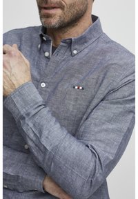 Camicia blu chiaro con bottoni e maniche lunghe, realizzata in tessuto testurizzato. Presenta un piccolo dettaglio ricamato con strisce rosse, bianche e blu navy.