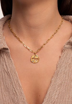 Collana di catena d'oro con un pendente circolare testurizzato recante la scritta "MAMA", adornato con pietre preziose colorate. Indossato su uno sfondo di tessuto neutro.