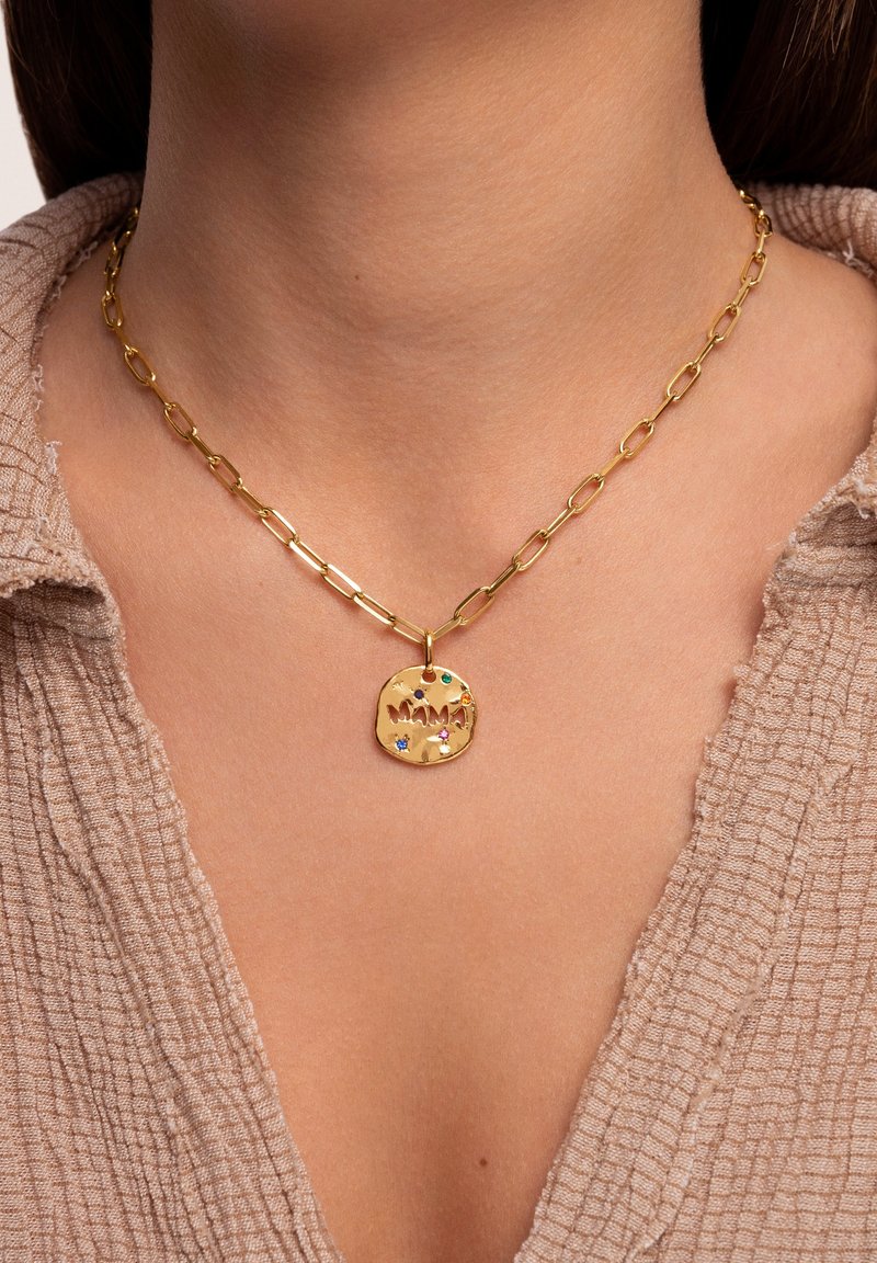 Collar de cadena de oro con un colgante circular texturizado que dice "MAMA", adornado con piedras preciosas de colores. Lleva sobre un fondo de tela de colores neutros.