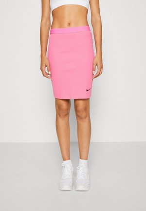Femme portant une jupe moulante rose de Nike, un soutien-gorge de sport blanc, des chaussettes blanches et des baskets blanches, debout devant un fond uni.