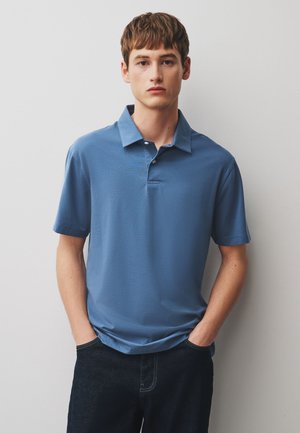 Jeune homme aux cheveux courts et bruns portant un polo bleu à manches courtes et un pantalon foncé, debout les mains dans les poches sur un fond uni.