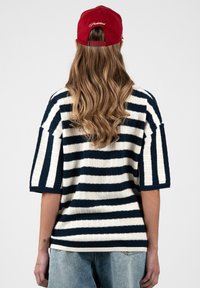 Persona con cabello largo y ondulado que lleva una gorra roja y un suéter de manga corta a rayas en blanco y negro, de pie de espaldas sobre un fondo liso.