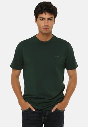 Giovane uomo con capelli ricci scuri che indossa una maglietta verde scuro a maniche corte e jeans scuri, in piedi con le mani in tasca.